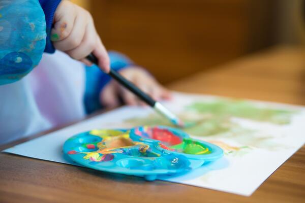 Bild vergrößern: Bild vergrößern: In Nahaufnahme ist eine Kinderhand zu sehen, die einen Pinsel hält und mit bunten Wasserfarben ein Bild malt.