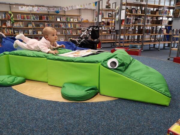 Bild vergrößern: Innenansicht der Stadtbücherei mit großem Stoffkrokodil als Sitzmöbel, auf dem ein Baby liegt
